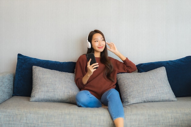 세로 아름 다운 젊은 아시아 여자는 거실 인테리어에 소파에 음악을 듣기 위해 헤드폰으로 스마트 휴대 전화를 사용