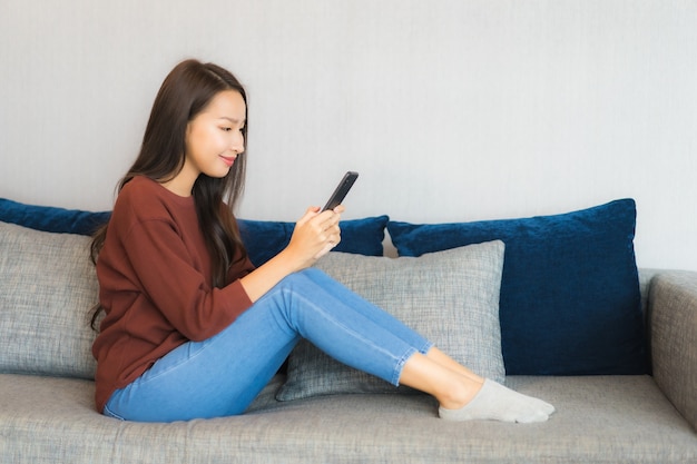 Телефон пользы женщины портрета красивой молодой азиатской умный на софе в интерьере живущей комнаты