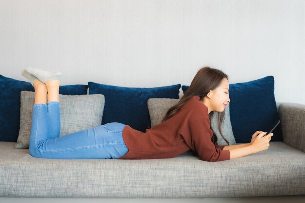 肖像画の美しい若いアジアの女性は、リビングルームのインテリアのソファーにスマートフォンを使用します。