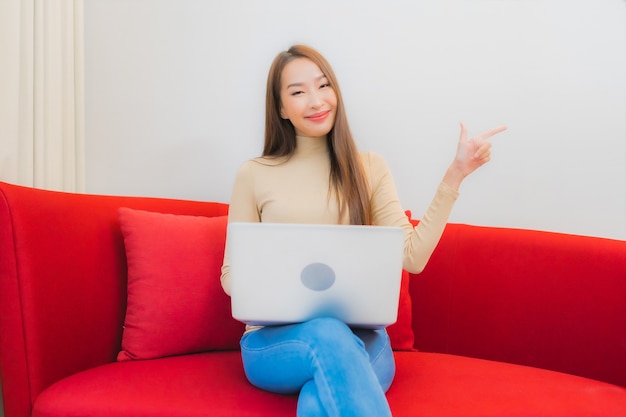 Портрет красивой молодой азиатской женщины использует компьтер-книжку компьютера на софе в интерьере живущей комнаты
