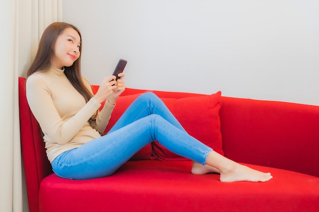 美しい若いアジアの女性の肖像画は、リビングルームのインテリアのソファでスマートフォンを使用しています
