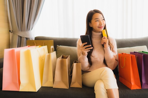 ラップトップまたはスマートモバイル携帯電話を使用してショッピングバッグの周りのソファでオンラインショッピングの美しい若いアジア女性の肖像画