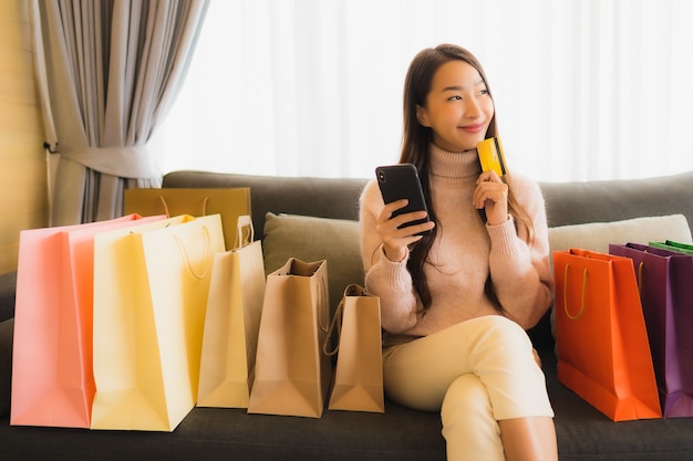 Женщина портрета красивая молодая азиатская используя компьтер-книжку или умный мобильный телефон для делать покупки онлайн на софе вокруг хозяйственной сумки