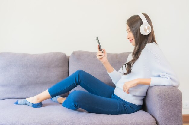 스마트 휴대폰과 헤드폰을 사용하여 거실에서 음악을 들을 수 있는 아름다운 젊은 아시아 여성의 초상화