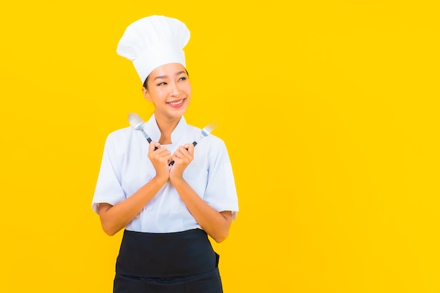초상 아름다운 젊은 아시아 여성은 노란색 외진 배경에 숟가락과 포크로 요리사 또는 요리 유니폼을 입고