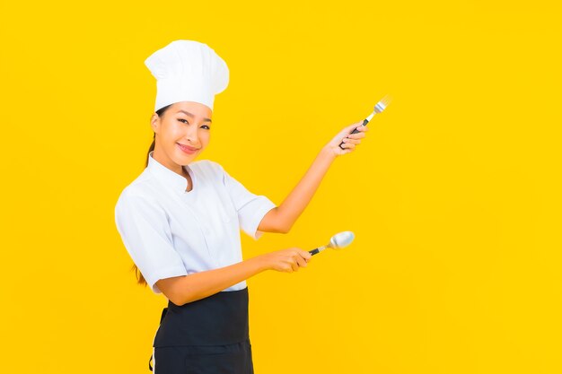 초상 아름다운 젊은 아시아 여성은 노란색 외진 배경에 숟가락과 포크로 요리사 또는 요리 유니폼을 입고