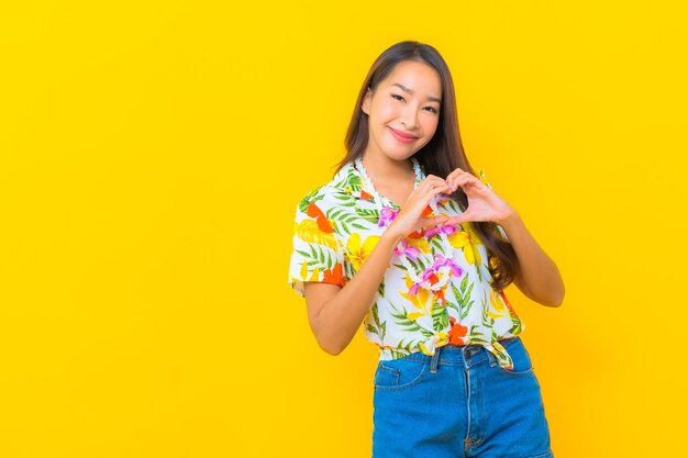 Portrait of beautiful young asian woman wearing colorful shirt and making heart sign on yellow wall