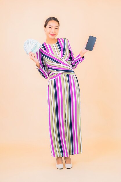 Женщина портрета красивая молодая азиатская с наличными деньгами или деньгами и мобильным умным телефоном на цвете