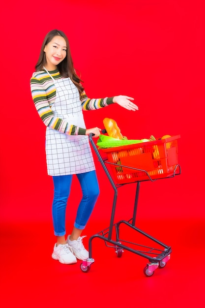 Foto gratuita bella giovane donna asiatica del ritratto con il cestino della drogheria dal supermercato sulla parete isolata rossa