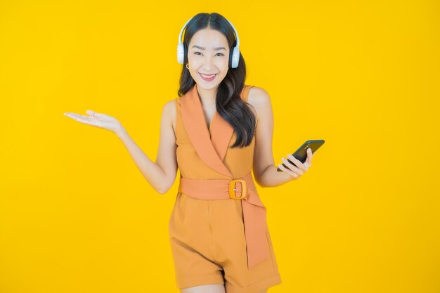 黄色の背景で音楽を聴くためのヘッドフォンとスマートフォンを持つ美しい若いアジアの女性の肖像画