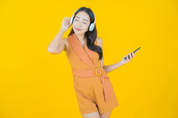 黄色の背景で音楽を聴くためのヘッドフォンとスマートフォンを持つ美しい若いアジアの女性の肖像画