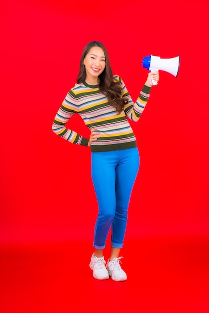 Женщина портрета красивая молодая азиатская с мегафоном для связи на красной стене