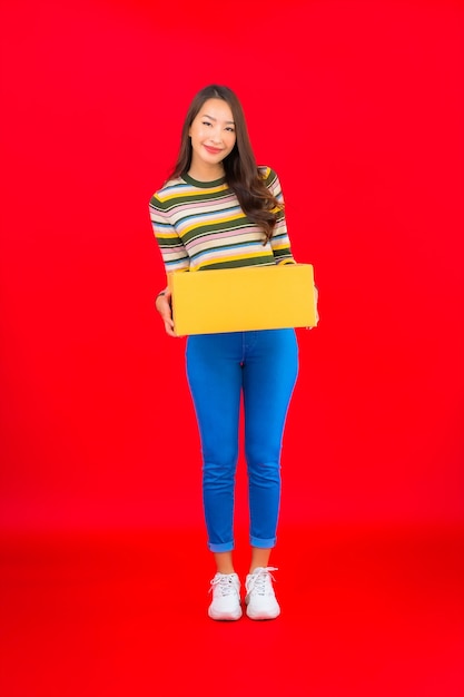 Женщина портрета красивая молодая азиатская с коробкой пакета на красной изолированной стене