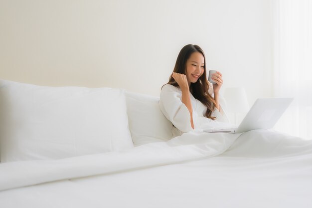 ベッドの上のコーヒーカップとコンピューターのラップトップを持つ美しい若いアジア女性の肖像画