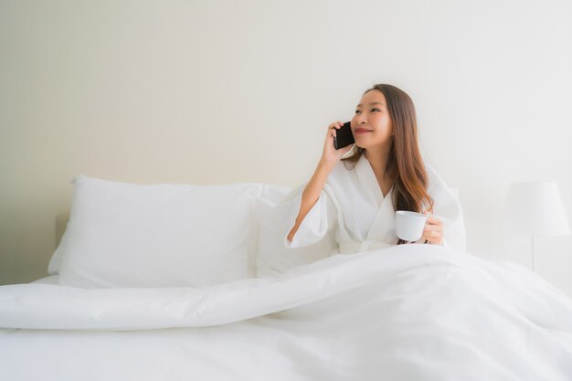 コーヒーカップとベッドの上の携帯電話を持つ美しい若いアジア女性の肖像画