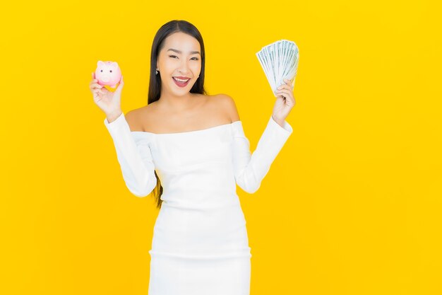 Портрет красивой молодой деловой азиатской женщины с большим количеством наличных денег и копилкой на желтой стене