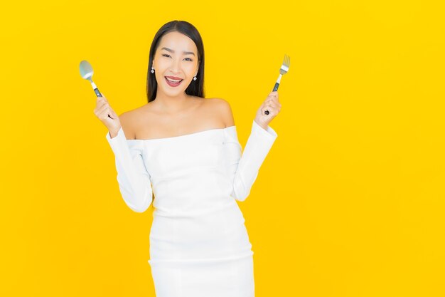 Портрет красивой молодой деловой азиатской женщины с ложкой и вилкой, готовой съесть еду на желтой стене