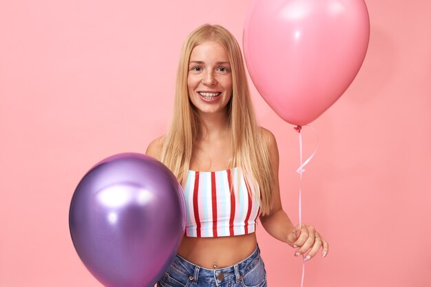 Portrait of beautiful young Caucasian woman with straight fair hair and braces wearing stylish summer top having fun, posing with festive decoration, holding two helium balloons