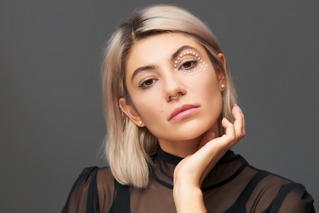 Portrait of beautiful young Caucasian woman with stylish haircut, nose ring and white crystals around one eye keeping hand on her face. Skin care, make up, cosmetics, cosmetology and beauty concept