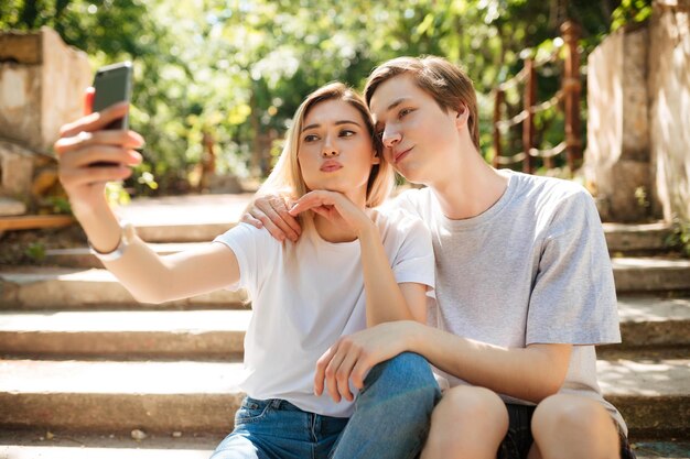 Portrait of beautiful young couple sitting on stairs in park and making selfie together Cool boy and pretty girl with blond hair looking in camera while taking photos on mobile phone frontal camera