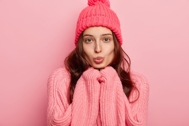 Portrait of beautiful young European woman keeps lips rounded, hands under chin, looks directly at camera, wears warm winter hat and sweater, poses over pink background.