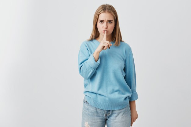 Portrait of beautiful young european woman with fair hair holding index finger at lips, asking to keep silence or not tell anyone her secret, saying "Shh", "Hush", "Tsss". Body language