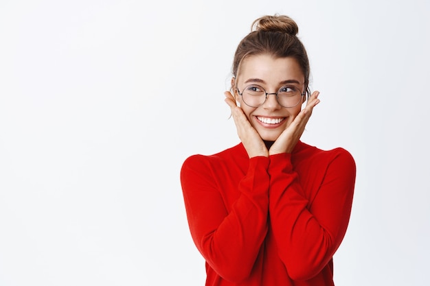 Free photo portrait of beautiful young female entrepreneur in glasses and red sweater, holding hands on cheeks and smiling, touching flushed face, standing over white wall