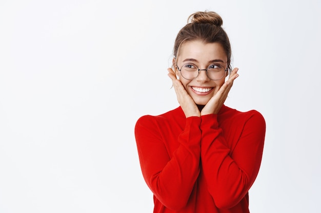 Ritratto di giovane e bella imprenditrice in occhiali e maglione rosso, tenendosi per mano sulle guance e sorridente, toccando il viso arrossato, in piedi sul muro bianco