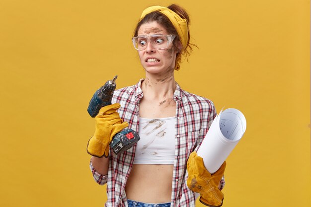 Portrait of beautiful young female wearing shirt, white top and protective eyewear holding drill and rolled paper looking with disgusting look at drilling machine isolated over yellow wall