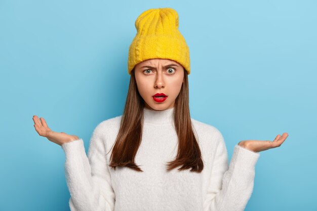 Portrait of beautiful young girl being questioned, spreads palms sideways, feels unawareness and doubt, wears red lipstick, wears yellow stylish hat, white jumper