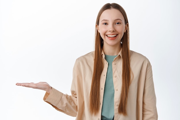 Portrait of beautiful young girl display in open hand, holding item on palm and smiling, demonstrate product, standing in casual clothes against white wall
