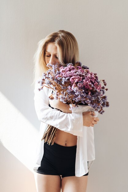 Portrait of a beautiful young girl in white shirt, black top and shorts, holding a big bouquet of dried flowers on gray