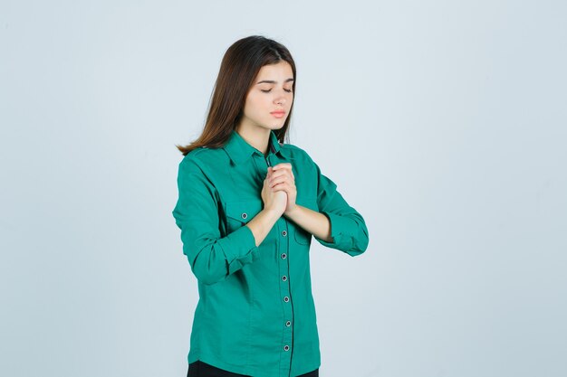 緑のシャツでジェスチャーを祈って手を握りしめ、希望に満ちた正面図を探している美しい若い女性の肖像画