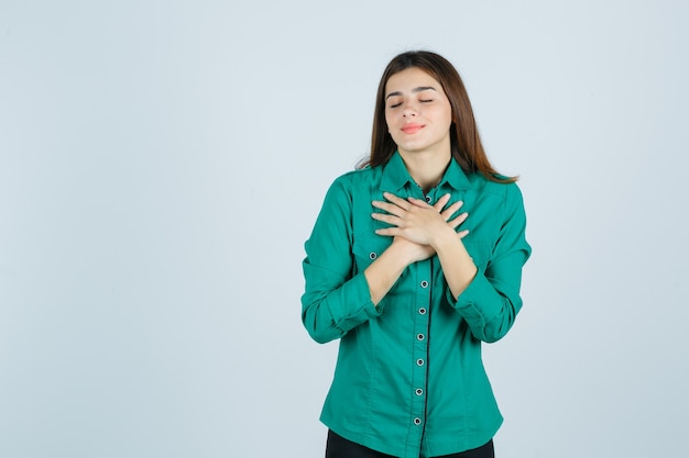 Portrait of beautiful young lady holding hands on chest in green shirt and looking delighted front view