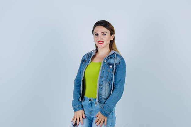 Portrait of beautiful young lady keeping palms on thighs in denim outfit and looking cheerful front view