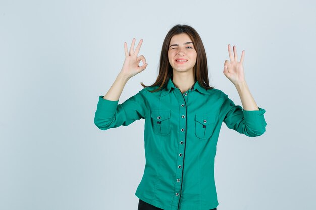 Портрет красивой молодой леди, показывающей жест ОК, подмигивая в зеленой рубашке и выглядя забавно