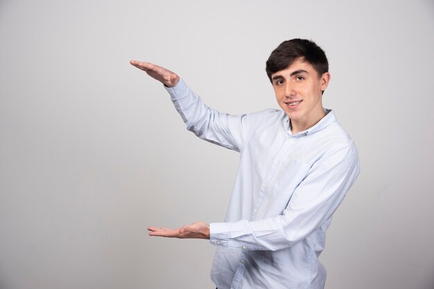 Portrait of beautiful young man showing size with hands against gray wall.