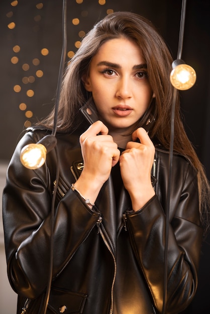 Портрет красивой молодой модели в черной кожаной куртке, позирующей возле ламп.