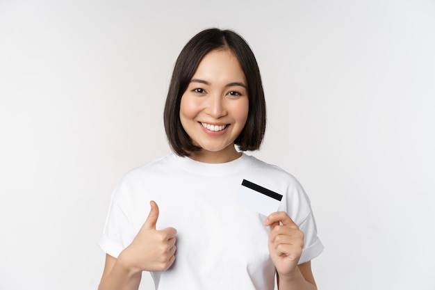 白い背景の上に立っている非接触型決済を推奨するクレジットカードと親指を示す美しい若い現代アジアの女性の肖像画