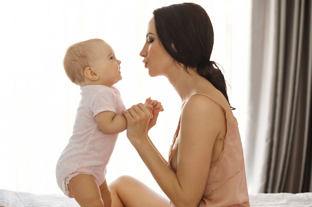 Portrait of beautiful young mom in sleepwear and her baby looking at each other sitting in bed Window surface.