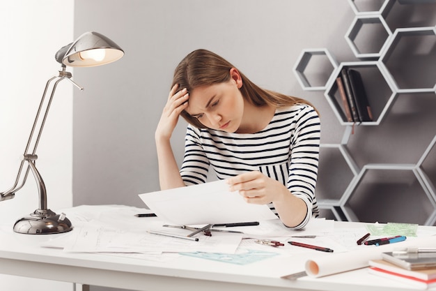 Portrait of beautiful young unhappy female engineer sitting at white table in cosy coworking space, looking at papers with upset expression being sad after finding mistake in blueprints.