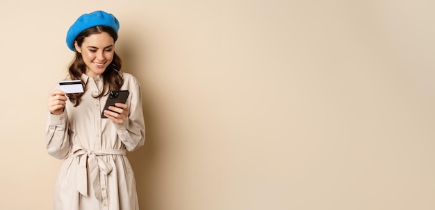 Foto gratuita ritratto di giovane e bella donna di 25 anni che utilizza l'app per lo shopping del telefono cellulare acquisto di consegna dell'ordine della carta di credito in piedi su sfondo beige