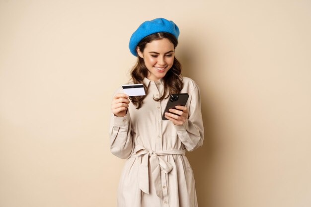 Portrait of beautiful young woman, 25 years old, using mobile phone shopping app, credit card, order delivery, purchase, standing over beige background