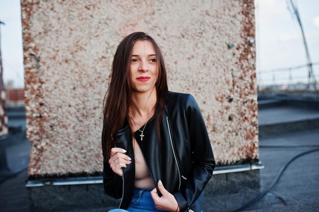 Portrait of a beautiful young woman in black leather jacket jeans and sneakers sitting on a rooftop