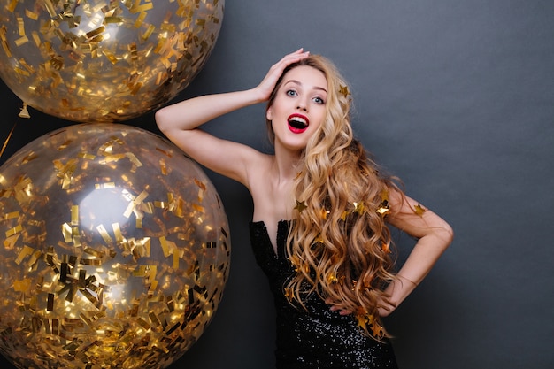 Portrait beautiful young woman in black luxury dress, with long curly blonde hair, red lips, with big balloons full with golden tinsels. Celebration, astonished, positivity.
