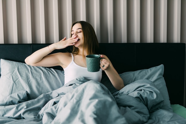 Портрет красивой молодой женщины, пьющей кофе на кровати
