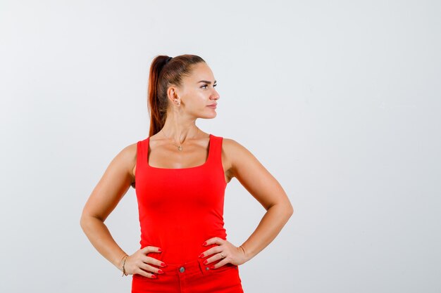 Portrait of beautiful young woman holding hands on waist, looking aside in red tank top, pants and looking confident front view