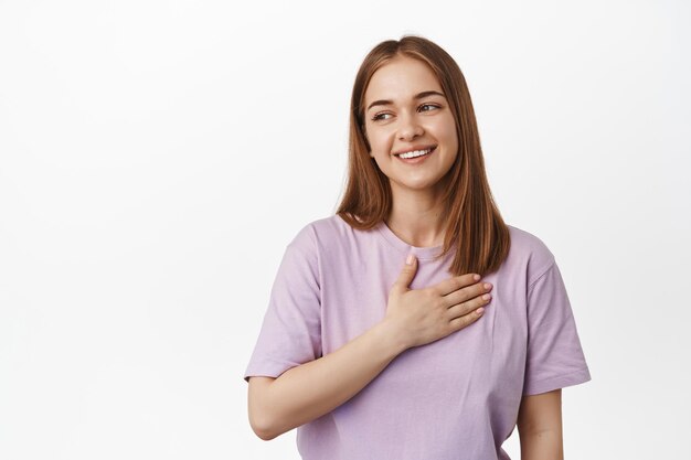 Portrait of beautiful young woman holds hand on heart, looking away at logo banner with pleased, flattered smile, laughing with genuine face expression, stands in t-shirt against white background.