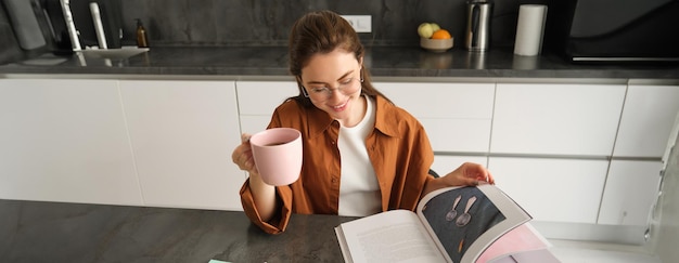 Portrait of beautiful young woman in kitchen sitting with a book flipping pages reading and drinking