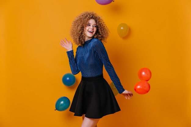 Portrait of beautiful young woman in mini skirt and blue sweater smiling on yellow background with colorful balloons